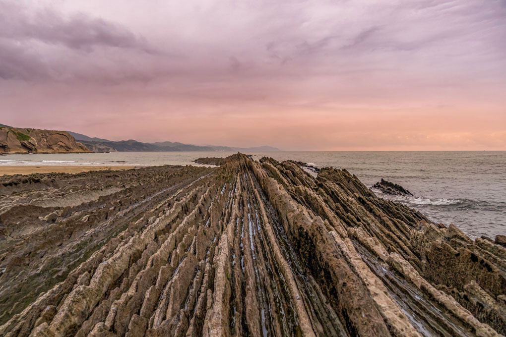 Costa rocosa con formaciones de estratos paralelos que se adentran en el mar, junto a una playa y montañas al fondo, bajo un cielo teñido de tonos rosados al atardecer.