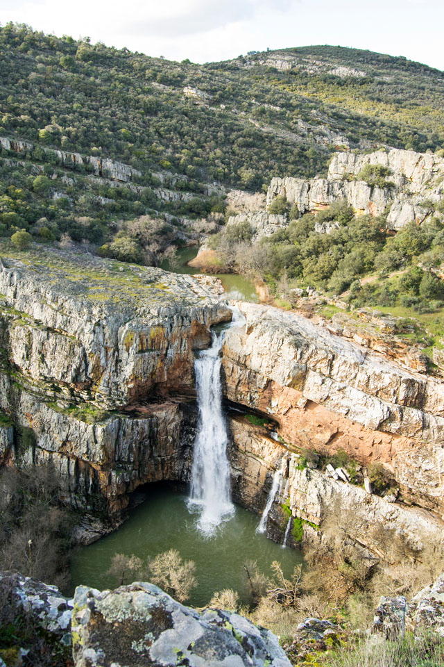 Salto de agua que cae entre grandes paredes de roca caliza hasta una poza rodeada de vegetación, en un paisaje montañoso de gran belleza natural.