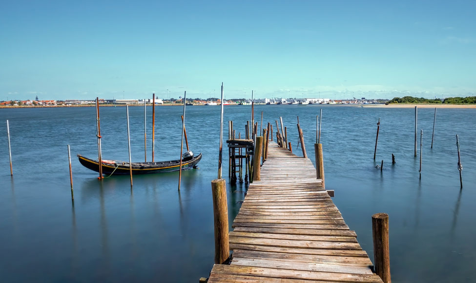 Embarcadero de madera sobre la ría de Aveiro, con una tradicional barca moliceira amarrada entre postes y el paisaje costero portugués al fondo.