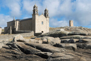 Iglesia de piedra de dos torres situada sobre un paisaje rocoso junto al mar, en Muxía. En primer plano destacan grandes formaciones de granito y, al fondo, un monumento de piedra, bajo un cielo azul con nubes.