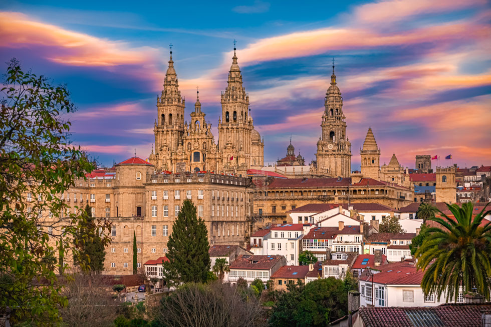 Vista panorámica de Santiago de Compostela al atardecer, con la Catedral de Santiago y sus torres destacando sobre el casco histórico, rodeadas de edificios tradicionales y tejados de la ciudad. Al fondo, un cielo de tonos azules, rosados y anaranjados.