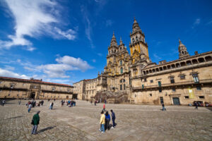 Plaza del Obradoiro de Santiago de Compostela, con la fachada barroca de la Catedral de Santiago al fondo y varios visitantes recorriendo la amplia plaza empedrada, bajo un cielo azul con nubes.