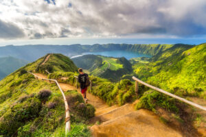 Sendero de montaña en São Miguel, en las Azores, con un excursionista recorriendo un paisaje volcánico cubierto de vegetación y vistas panorámicas sobre lagos y el océano Atlántico.