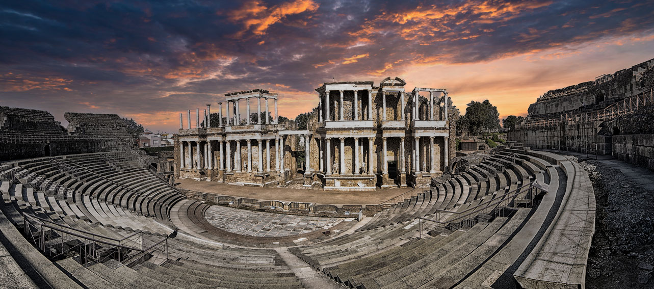 Teatro romano de Mérida al atardecer, con sus gradas semicirculares y el escenario monumental de columnas y esculturas bajo un cielo cubierto de nubes.