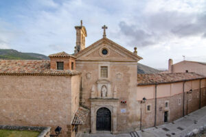 Edificio histórico de piedra en Cuenca, con una fachada de estilo religioso, portada con escultura y cruz, rodeado de tejados tradicionales y montañas.