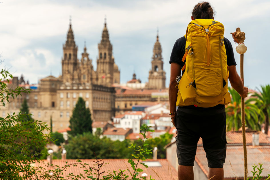 Peregrino con mochila y bastón contempla desde una colina las torres de la Catedral de Santiago de Compostela, rodeada por los tejados de la ciudad.