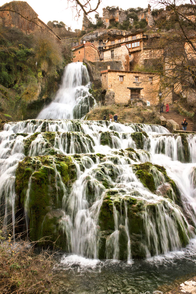 Cascada de Pedrosa de Tobalina, en Las Merindades (Burgos), con varios saltos de agua entre rocas cubiertas de musgo y antiguas construcciones de piedra y madera al fondo.