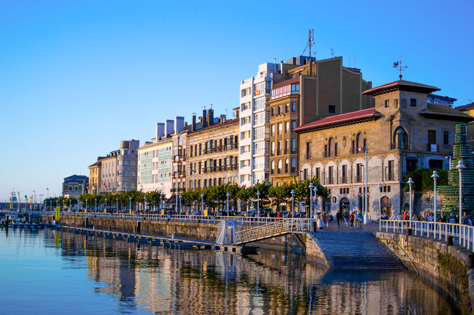Puerto deportivo de Gijón junto a Cimadevilla, con edificios históricos y embarcaciones reflejados en el agua.