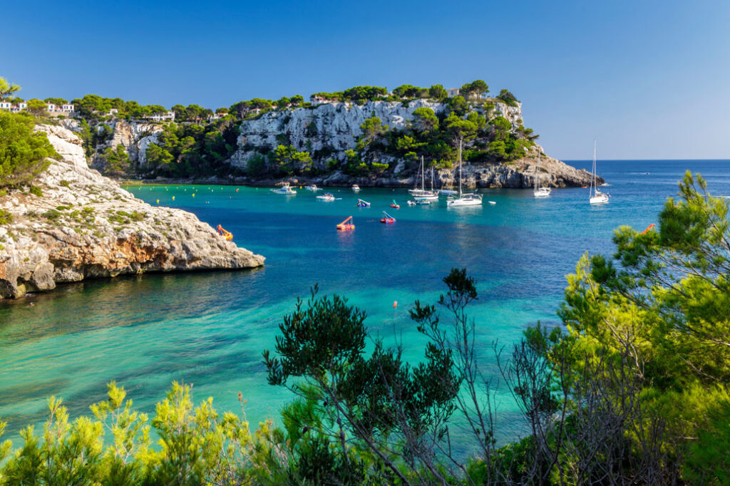 Cala de aguas turquesas en Menorca, rodeada de acantilados de roca blanca, pinos y vegetación mediterránea, con varias embarcaciones fondeadas en el mar.