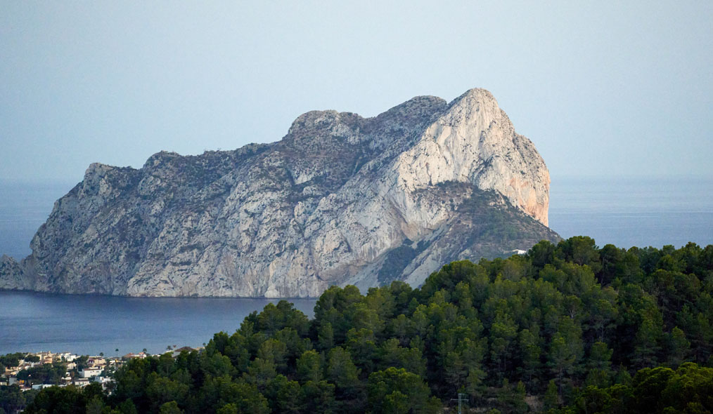 Vista del Peñón de Ifach, imponente formación rocosa que se alza frente al Mediterráneo, con el mar y el paisaje boscoso y urbano de la costa al fondo.