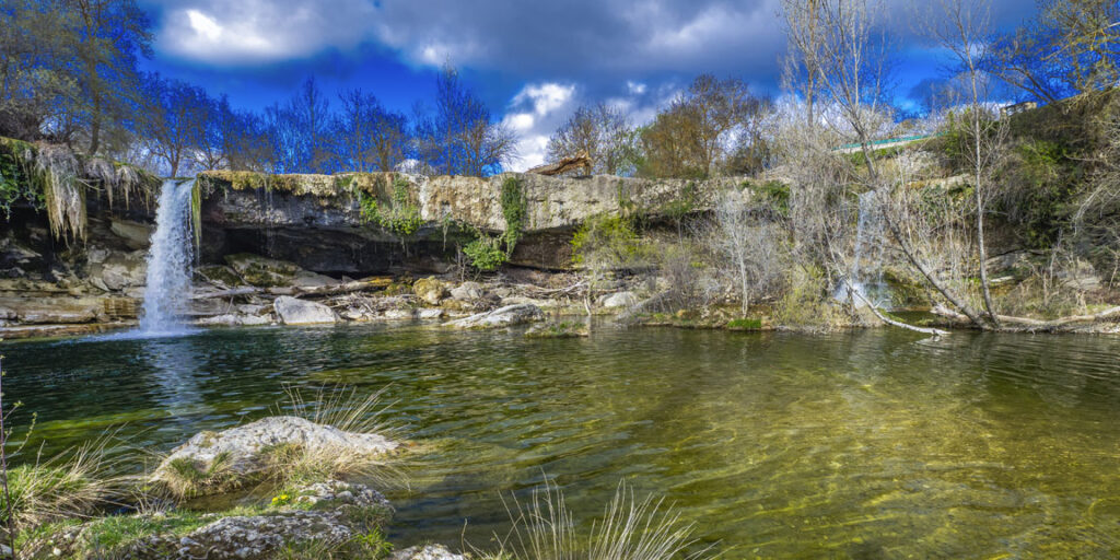 Paisaje de Las Merindades, en Burgos, con una cascada que cae sobre una poza de aguas cristalinas junto a un antiguo puente de piedra, rodeada de vegetación y árboles.
