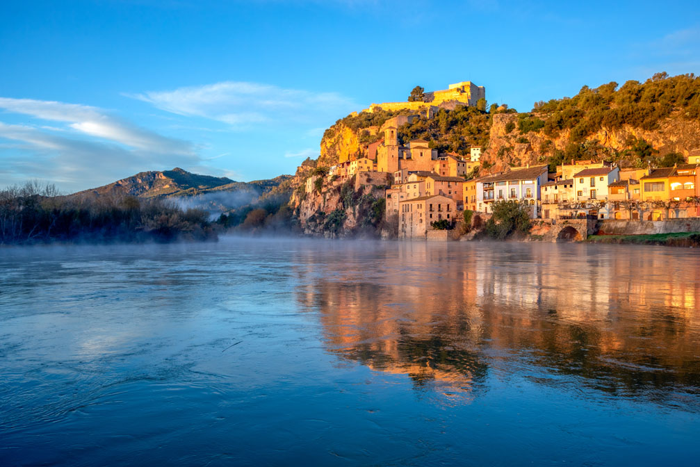 Vista de Miravet al atardecer, con el río Ebro en primer plano reflejando las casas del pueblo y la luz dorada. El casco histórico se extiende por la ladera rocosa, coronada por el castillo templario, rodeado de vegetación.