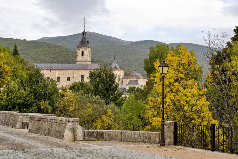 Fotografía del Monasterio de El Paular, en Rascafría, rodeado de abundante vegetación y con las montañas al fondo. En primer plano se observa un puente de piedra y una farola, mientras los árboles presentan tonos verdes y amarillos propios del otoño.