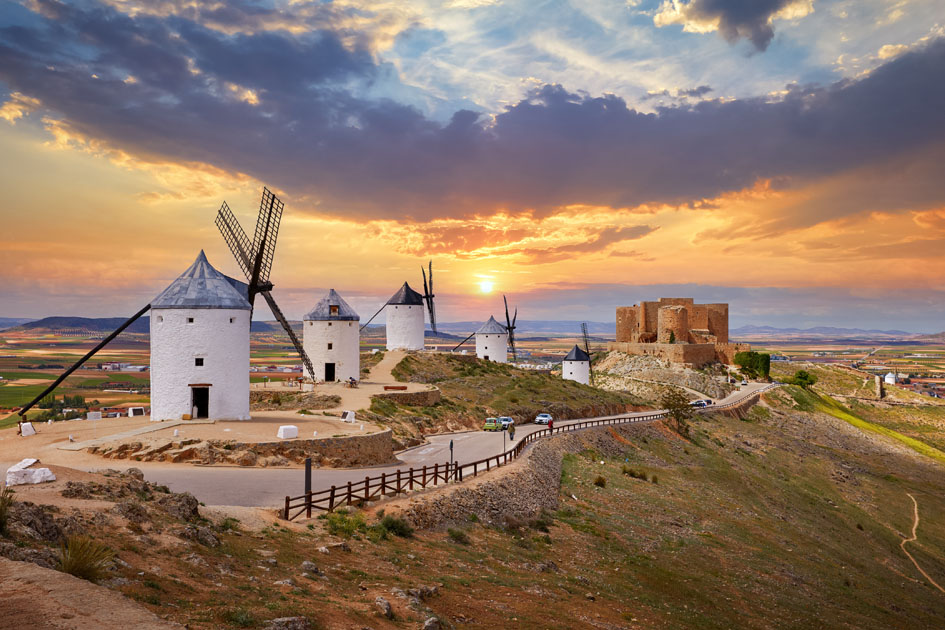 Paisaje manchego al atardecer con varios molinos de viento tradicionales sobre una colina y los restos de un castillo al fondo, rodeados de extensos campos de cultivo.
