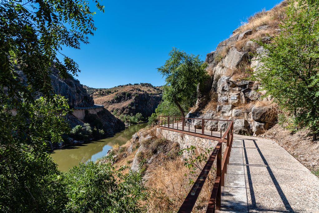 Senda fluvial junto al río Tajo, con un camino acondicionado entre paredes rocosas, vegetación de ribera y un paisaje de monte bajo bajo un cielo despejado.