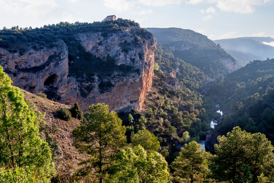 Paisaje de la Serranía de Cuenca, con imponentes paredes rocosas sobre un valle boscoso atravesado por un río, rodeado de pinares y vegetación mediterránea.