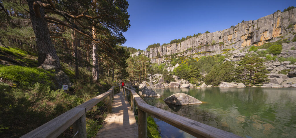 Paisaje de la Laguna Negra de Soria, con una pasarela de madera junto al agua, rodeada de pinares, rocas y paredes de piedra bajo un cielo despejado.